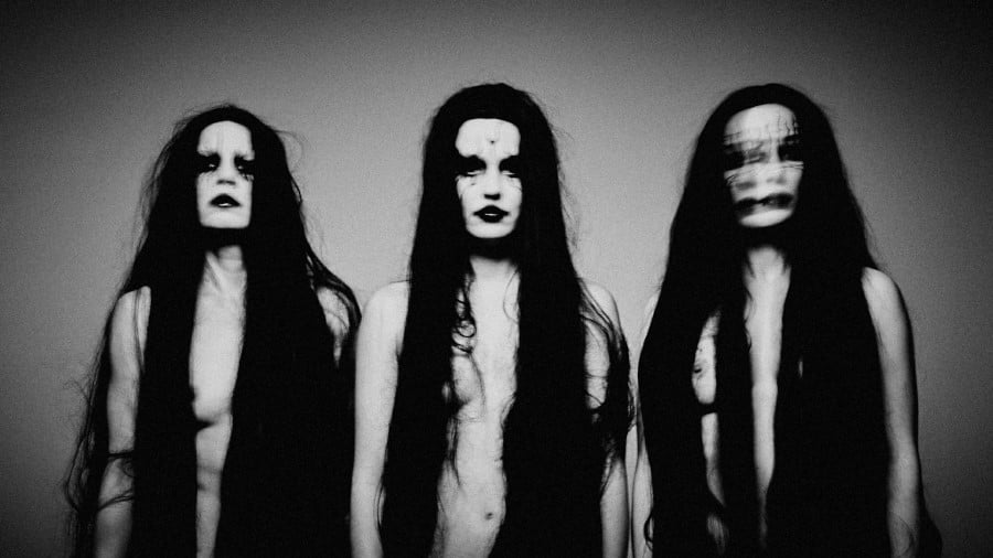 A black and white photo of three women with black lipstick and eye makeup, who appear naked, with long black hair (mostly covering their breasts.