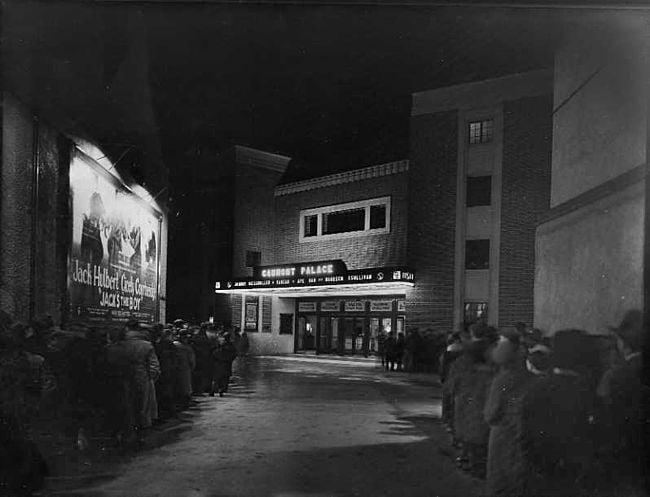 The courtyard leading to the main entrance to the Gaumont Palace, at night. Two queues are formed, snaking either side of the courtyard. The canopy over the foyer is lit up.
