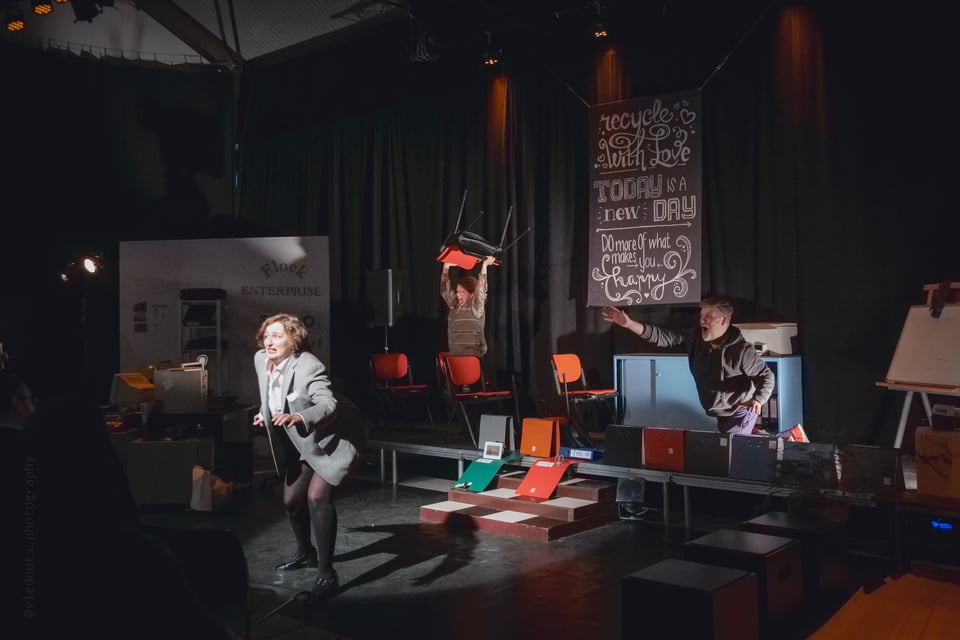 a set of an office with an actress running, an actress holding a chair above her head and an actor reaching out from behind a wall of folders