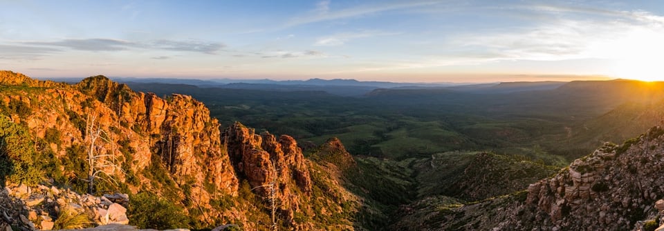 Sunset from the edge of the Mogollon Rim, home of the Four Corners potato. In the foreground rugged light brown rocks studded with sparse plants. The view stretches away to a distant horizon with the ball of the sun at the far right.