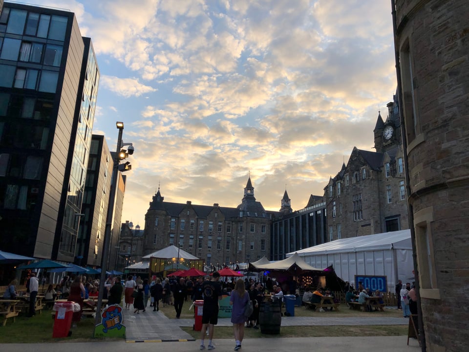 A green at sunset. Tall, Gothic revival buildings in a sandy grey along the back and right-hand side; slick modern buildings, deep grey along the left. Tents and parasols of all sorts, with people milling around.