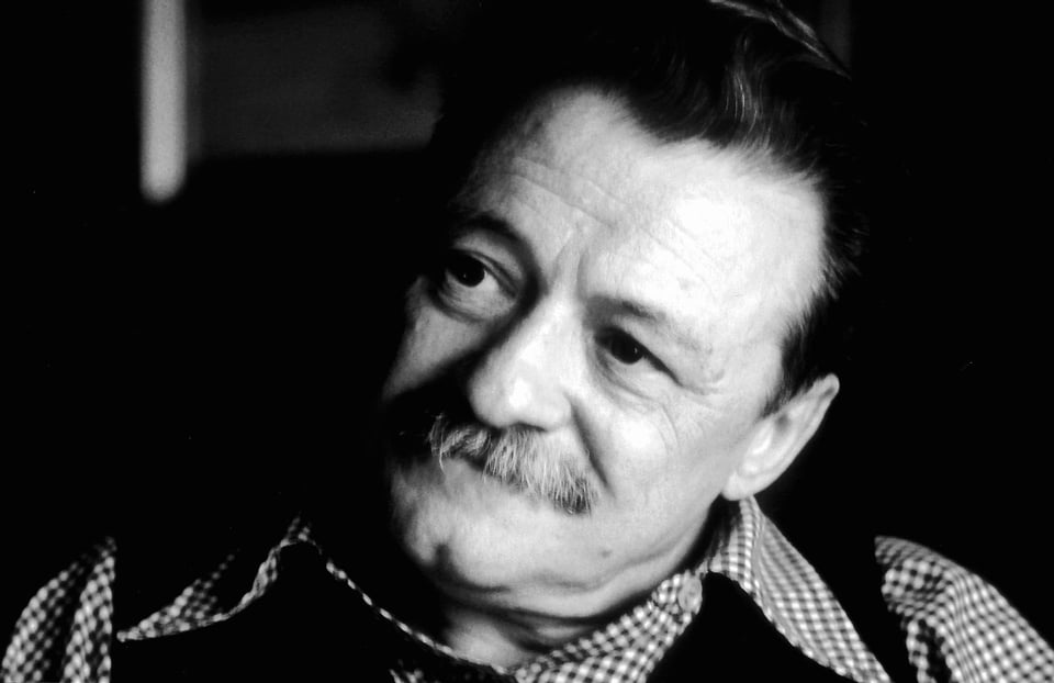 A black-and-white photography of Mario Benedetti, the poet whose work I translate in this post. He is a white man with slicked-back dark hair, dark eyes, thin graying eyebrows and a bushy gray mustache, wearing a checkered open-necked shirt and, I believe, some sort of sweater vest. He looks to his left, away from the camera, with the shadow of a wistful smile playing upon his half-hidden lips.