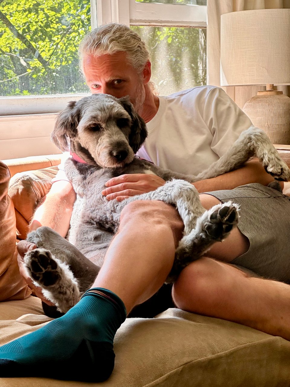 photo of white man in shorts and white T-shirt curled up with a big gray Aussiedoodle on a light brown leather sofa, with sunlight streaming from a window behind them.