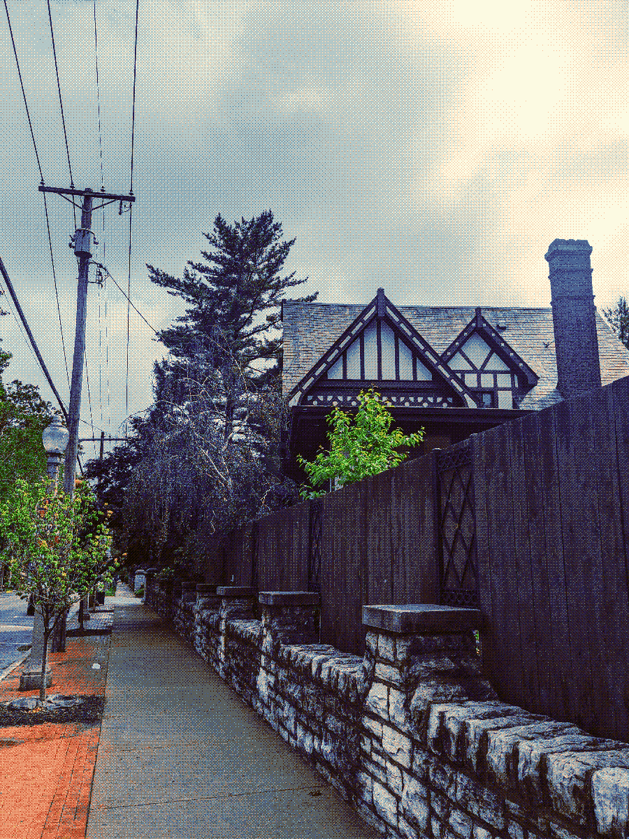 An old stone wall and a high wooden fence running along a sidewalk. Treetops and a house built in old-fashioned Western European style peek out over the fence into a cloudy sky.