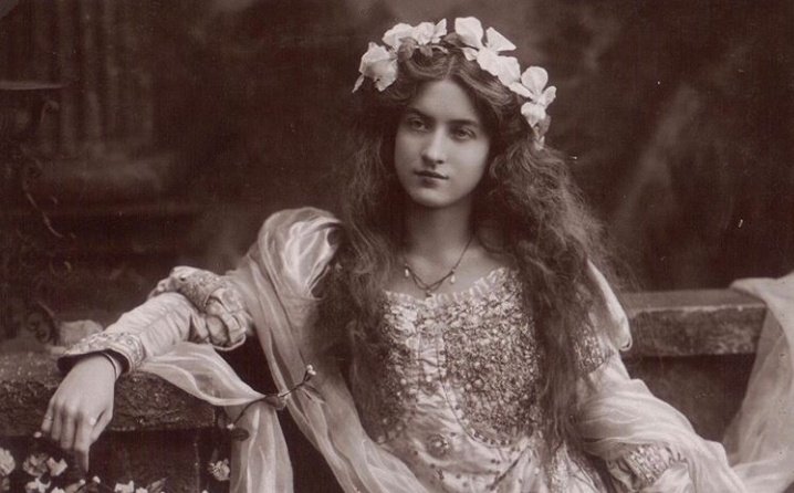 A photo of actress Maud Feely sitting on a stone bench in a flower crown and flowy, beaded and embroidered dress.