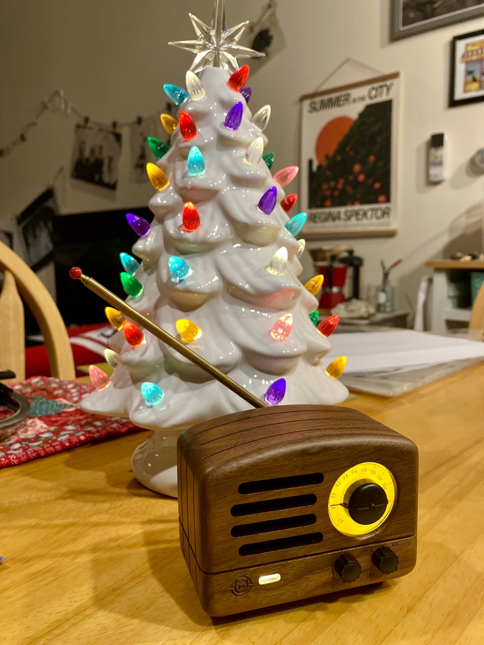 photo of a small Bluetooth speaker shaped like an old-fashioned wood-panel radio; behind it is a white ceramic Christmas tree with colored lights.