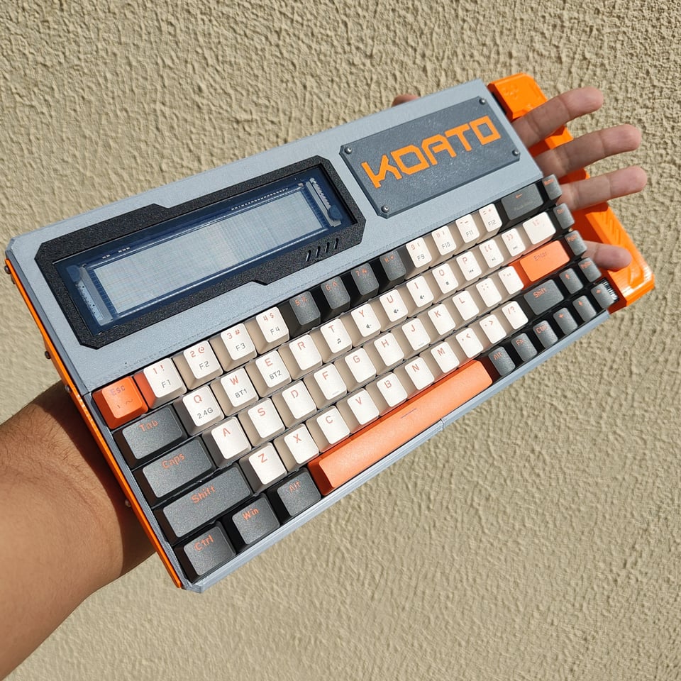 A cyberdeck computer that is about 18 inches long and 8 inches high; it has a white-black-orange "clicky" keyboard built in, and a small screen on the upper left corner. It has an orange handle on the far right side through which the person holding it -- mounted on their left forearm -- is sticking their fingers through, showing how you'd grip it securely