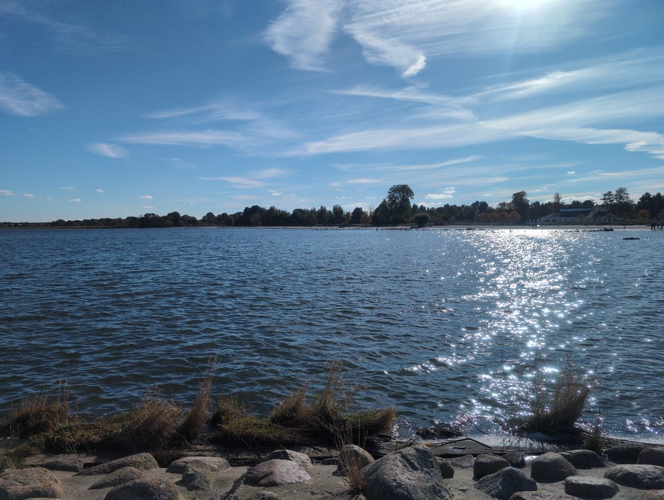 Picture of a seascape. There are rocks in the foreground leading to the water's edge. The water is dark blue and takes up the bottom half of the frame. There is sunlight reflecting on the right side of the water. At the far edge of the water is a row of trees in silhouette with a sandy shoreline slightly visible. Above the trees is a lighter blue sky with wispy clouds.