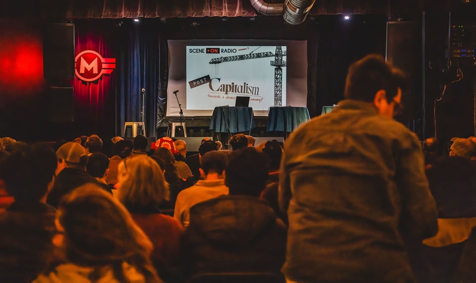 Image of audience gathering at Motorco -- on stage, screen showing Scene on Radio: Capitalism logo