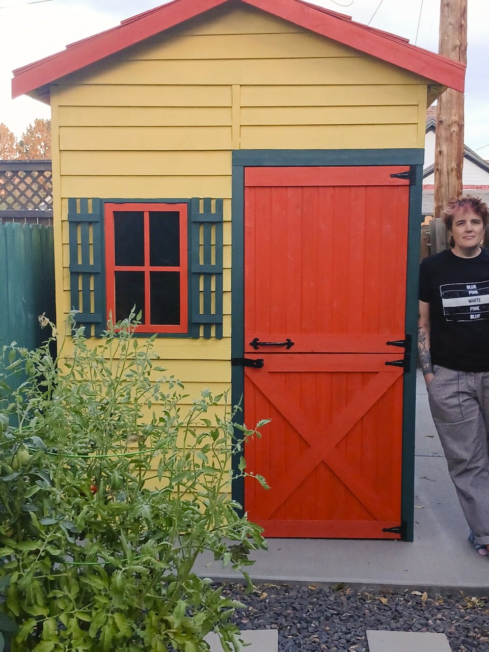 Me in striped trousers and a black-and-white t-shirt leaning against a small shed in our narrow back yard. Tomatoes are growing in front, and the shed is painted yellow, with an orange door and deep green-blue trim. It's a douch door, split top and bottom, with a small window next to it.