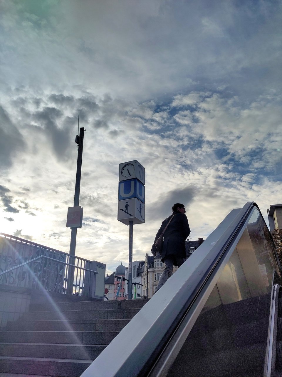 Blick den Handlauf einer Rolltreppe entlang aufwärts, bis hinauf in einen locker bewölkten Himmel.