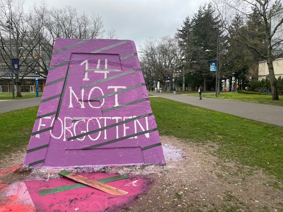 A large concrete cairn sits on a campus lawn. The concrete is painted mauve, with the words '14 NOT FORGOTTEN'. Buildings, walkways, and trees are visible in the background.