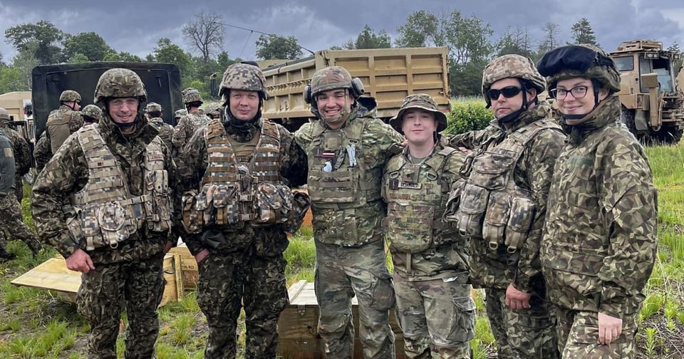A group of six soldiers in full camouflage gear pose together on a grassy field. Military vehicles and equipment are visible behind them under an overcast sky.