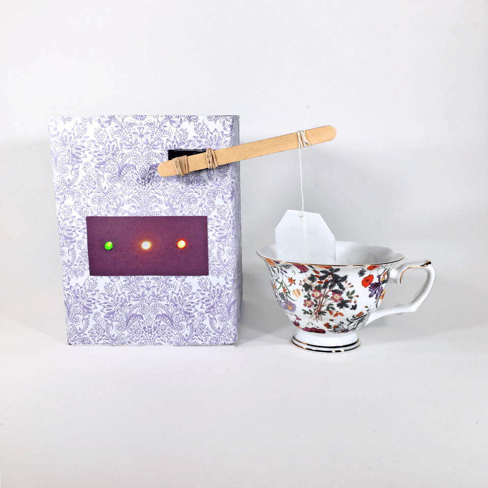 A picture of a whimsical DIY tea-brewing gadget. It is wrapped in lavender damask paper and has three three LED lights -- green, yellow, and red -- and a popsicle stick arm, attached with rubber bands to the top of the device, holding the bag by its string and dipping it into an elegant patterned teacup. It looks pretty funny and sweet
