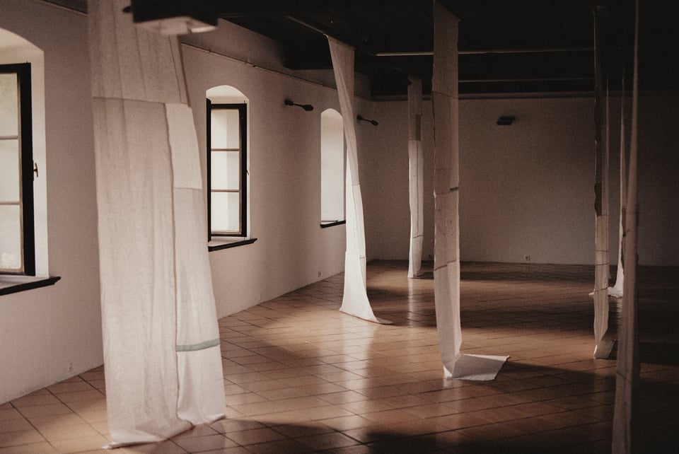 [A wide corridor with a tile floor and lots of light. Panels of stitched-together white fabric hand from the ceiling to the floor, scattered throughout the space.]