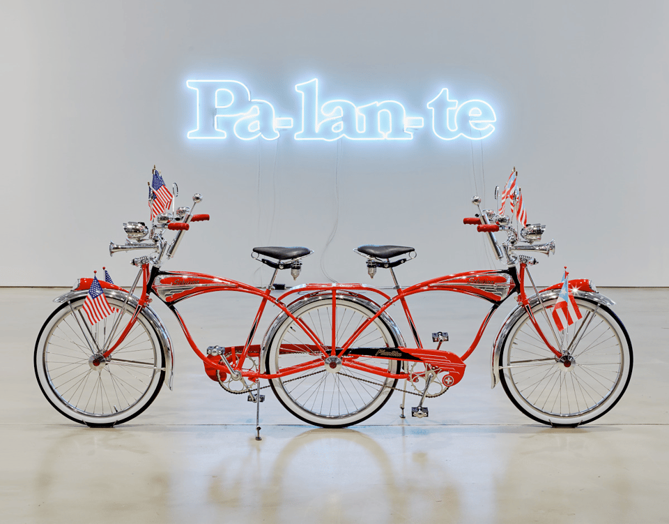 Two cherry-red Schwinn bicycles, decked out with horns and American and Puerto Rican flags, are joined to form one three-wheeled, double-headed creature. The word pa'lante, Spanish slang meaning "forward," glows above.