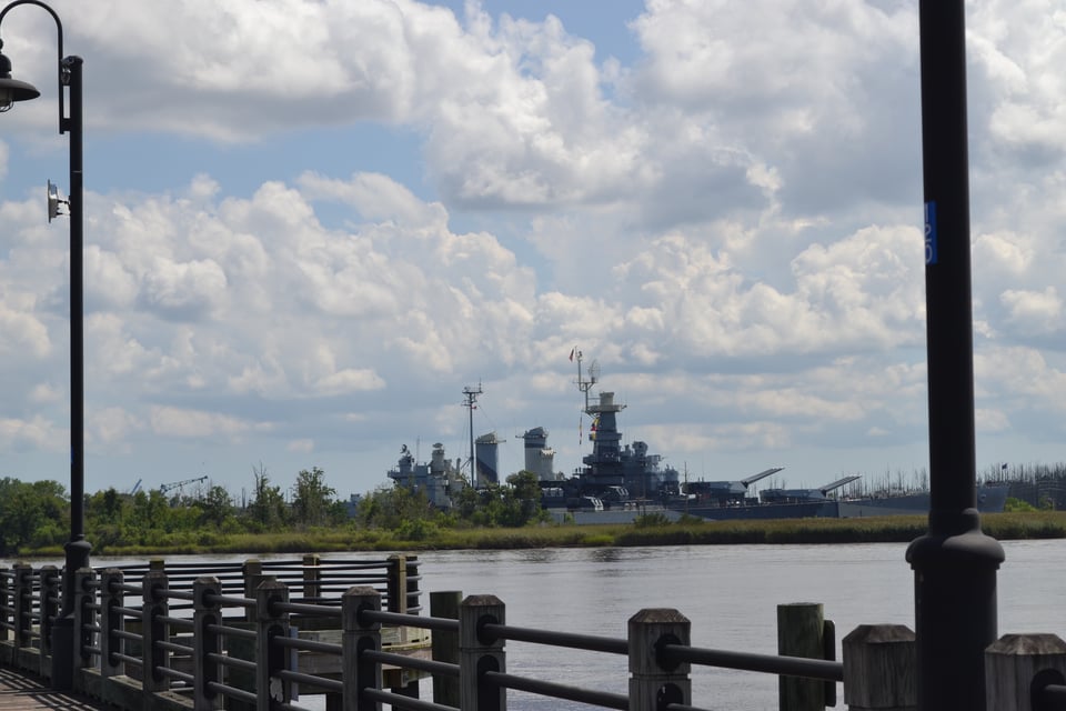 A wide river viewed from a dock with a railing. On the far side of the river is a huge warship with lots of guns.