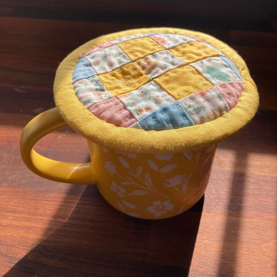 A yellow and white. floral mug of tea rests on a dark wooden desk in the morning light; it has a colorful patchwork fabric lid on top of it to keep the heat from escaping.