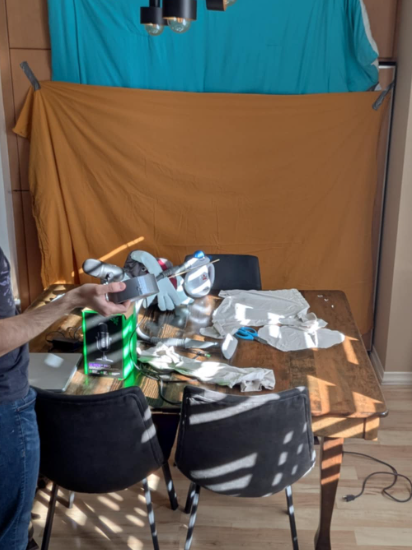 Kurt stands partially offscreen, holding a roll of tape. In the back, two sheets are taped to the wall. On the table, various craft supplies, recording materials, and Herman the puppet are strewn about.