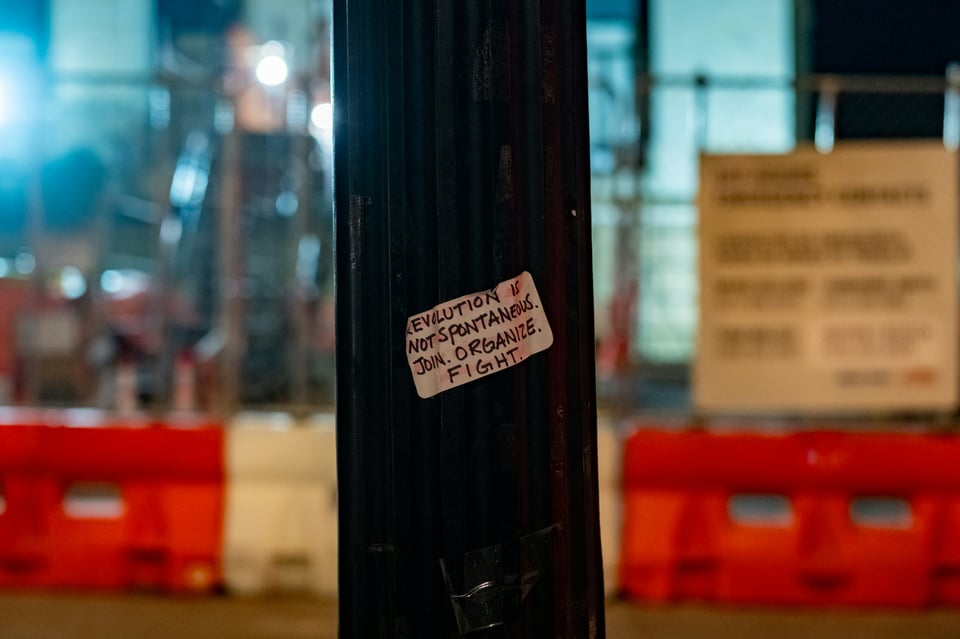In focus is a pole in the foreground, with a white, rectangular sticker with black writing that reads "Revolution is not spontaneous. Join. Organize. Fight." In the background, out of focus, is a white and orange construction barrier with a chain link fence.