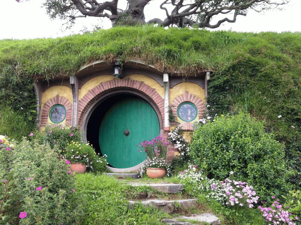 A photo of a house built into a hill. The house has a large green, round door, with red and yellow decorations around the edges, and a small round, stained glass window to the right and left of the door. There are several steps leading up to it, and the hill into which the house is built is covered in green grass. In the background we see a gnarled tree with no leaves reaching towards the sky