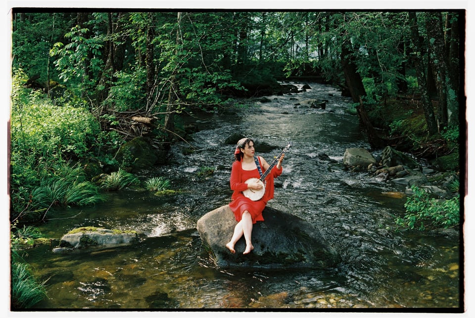 A woman in a red dress sits on a rock in a stream, playing a banjo.