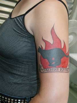 A profile of my torso, with my left arm foregrounded. On my arm is a fresh tattoo of a black cat running with a fire in the background. Underneath a banner reads CONCORDIA DISCORS.
