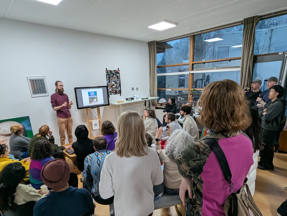 A group of people cluster around a TV in an expanding ring of couches and benches. Some at the back are standing, and listening to a speaker in a plum shirt and orange wine pants.