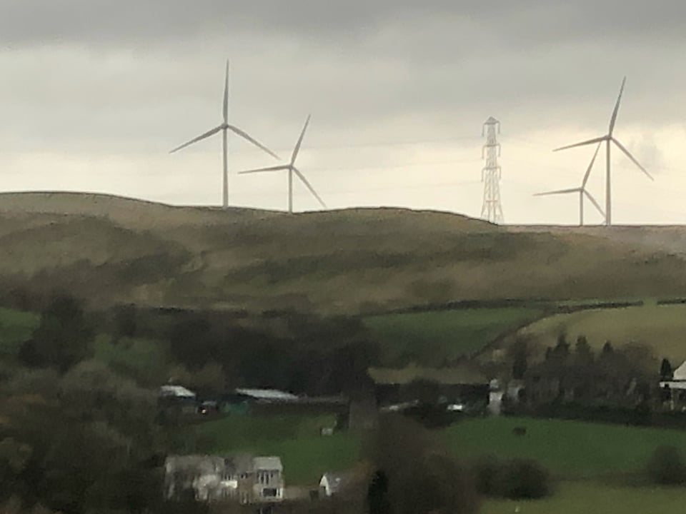 A grass-covered ridge with grey clouds above. Poking about the ridge are four three-bladed white wind turbines.