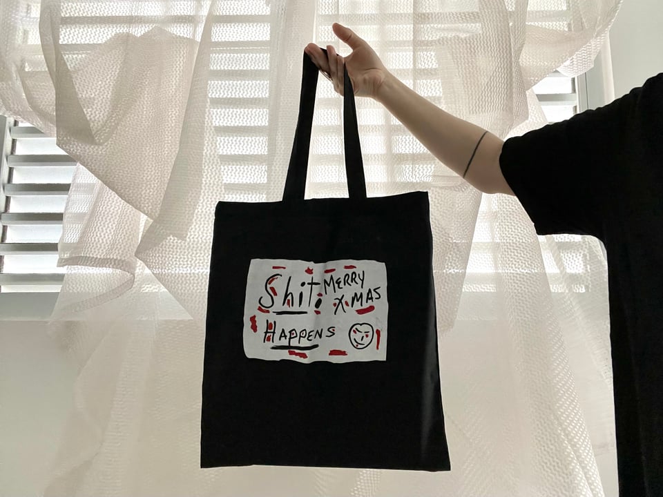 Photo of a black tote with a black, white, and red recreation of a handwritten sign that says “Shit happens, Merry Xmas”