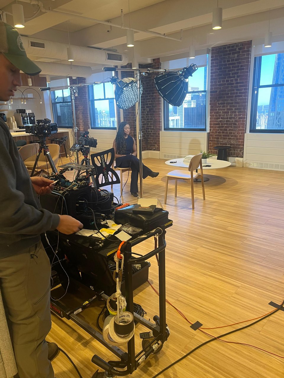 Photo of me sitting in a TV studio on a wooden chair, facing a second empty wooden chart. There are lights and cameras on me. There's a man to the side looking at filming equipment.