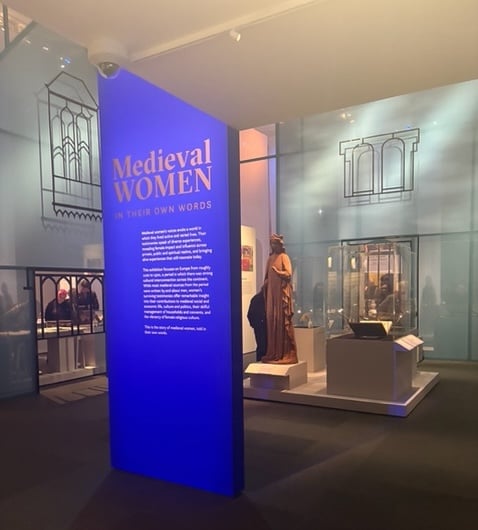 photo of the beginning of the Medieval Women exhibition at the British Library in London. a blue sign welcomes you, while there are objects in the background