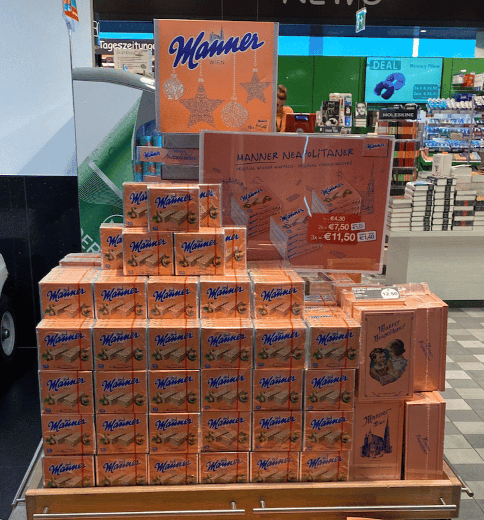 A tall display of small boxes of cookies in an airport shop. The boxes are all a pink-orange color, with blue writing that says Manner, and photos of wafer cookies and hazelnuts. There is a sign with prices in euros (4.50, 7.50 and 11.50)