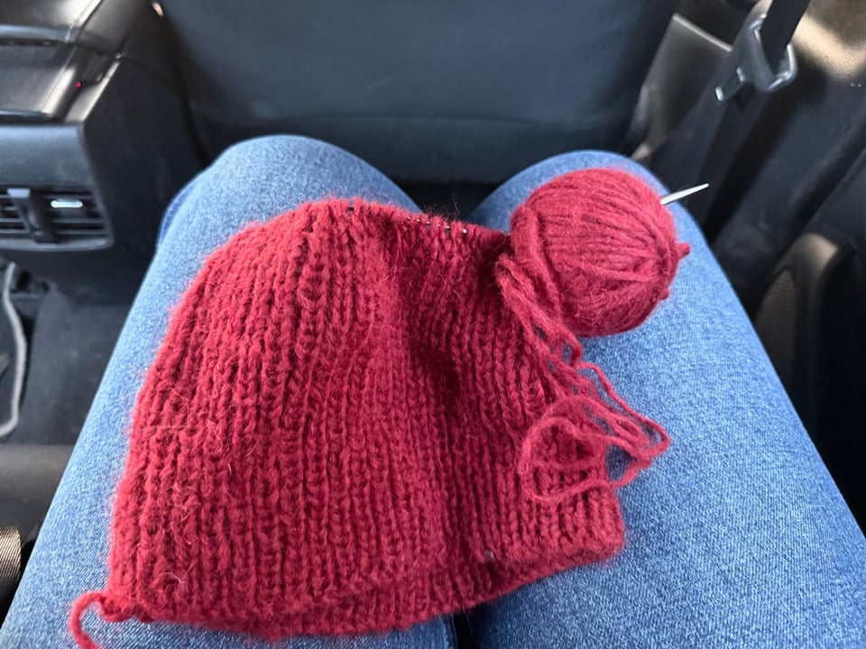 A person wearing a pair of jeans sits in an airplane. One the legs are the beginnings of a dark red knit hat.