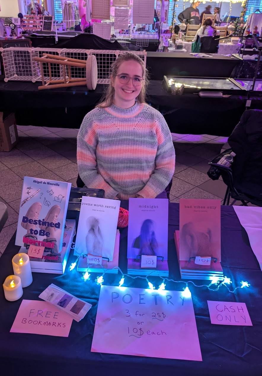 A photo of Abigail sitting at a table with a black tablecloth. Her books are displayed along with several signs with pricing and free bookmarks. The table is lit up with string snowflake lights and fake candles.