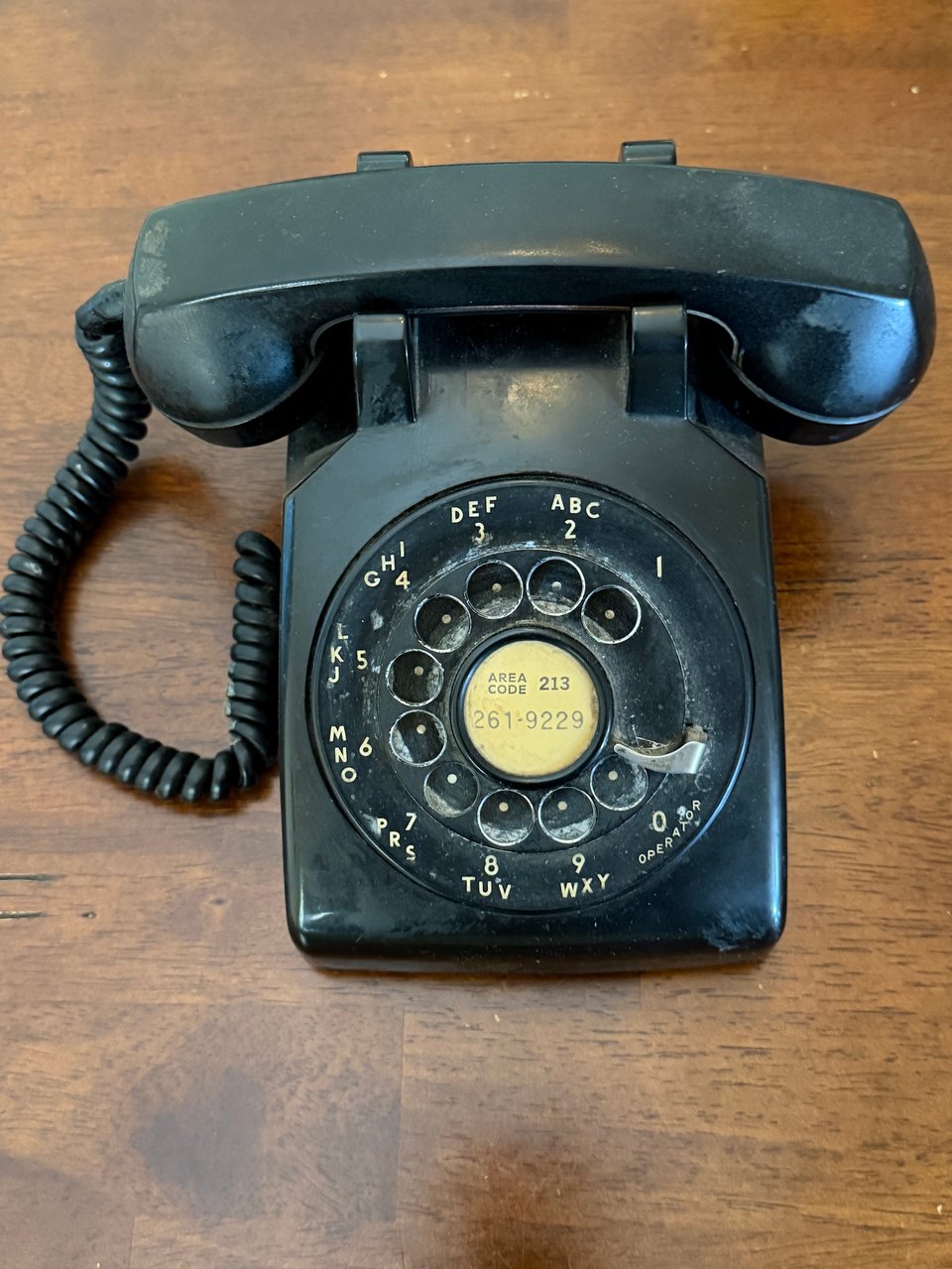 A weathered black rotary phone that belonged to my grandmother. Her old phone number is typed in the middle of the phone: AREA CODE 213 and underneath 261-9229. The phone sits on a wood table.