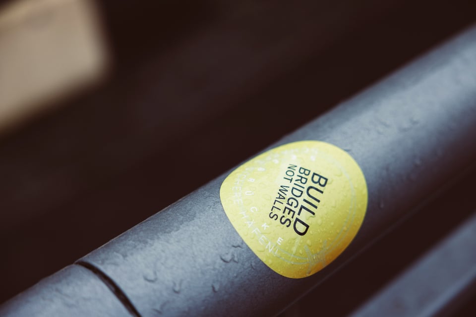 A circular yellow sticker with black text is upside down on a black railing. It reads "Build Bridges Not Walls." The railing and sticker have some drops of water on them like it has rained or is raining.