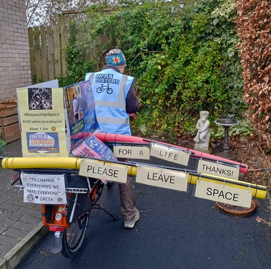 A woman stands with her decorated cargo bike - signs state '#RespectHerSpace ALSO when on a bicycle! Allow 1.5m', 'To change everything we need everyone - Greta', and 'Please leave space for a life thanks!'. The latter is hung from two cargo-rail mounted foam pool noodles which create an artificial width to the bike to encourage passing further out in the road.