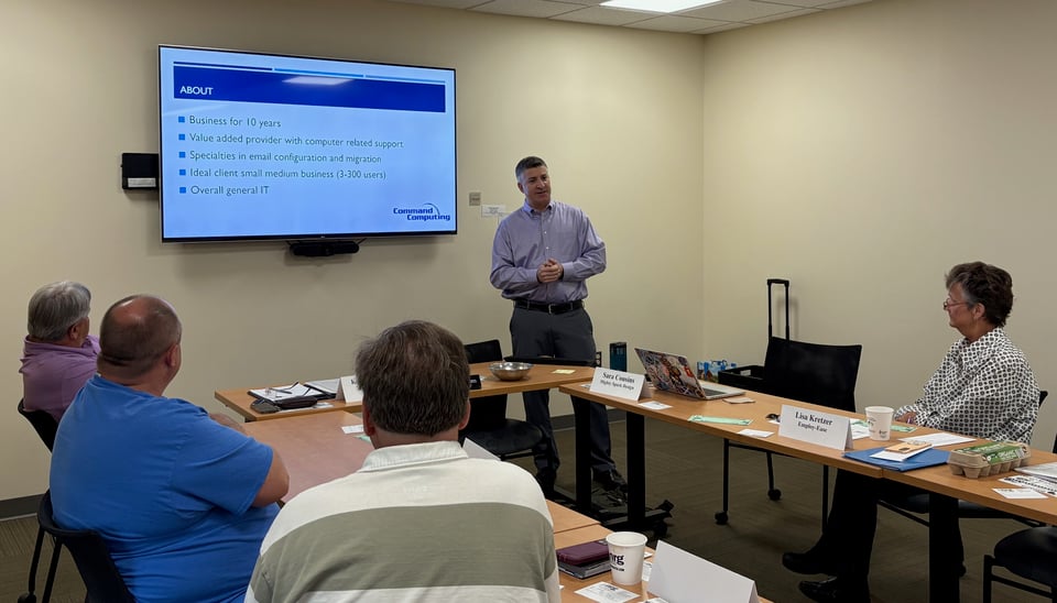 Rich stands in front of a group of people seated around the room next to his presentation displayed on a wall-mounted TV.