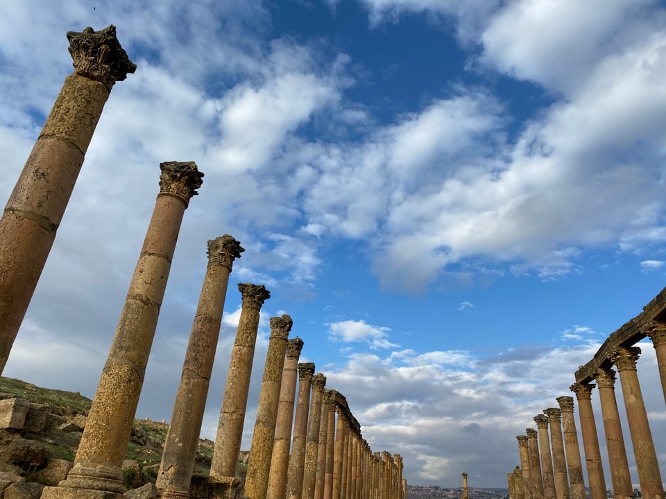 Photograph of two long rows of ancient Roman columns extending in parallel into the distance under a partly cloudy blue sky. Columns are weathered stone with Corinthian capitals