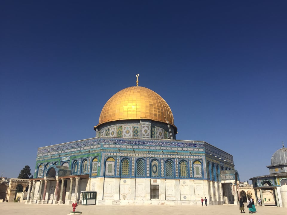 Photograph of Dome of the Rock showcasing its iconic golden dome and intricate blue and green tilework on walls. Clear blue sky and a few people near the structure
