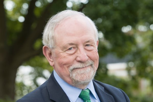 Portrait of JMC Prof. Norm Graham with white hair and a beard, wearing a suit and green tie outdoors.