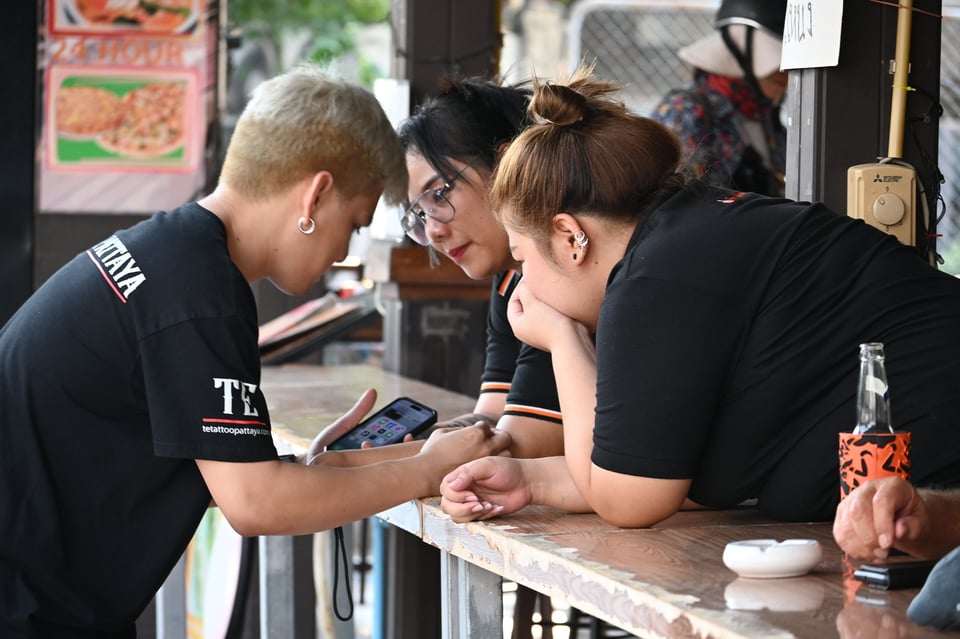 DSC_3942: Three friends lean on a counter, intently looking at a phone together while chatting in an outdoor market setting.
https://www.flickr.com/photos/pattayapatrol/53475695564/in/photolist-2ryDPKU-2qx7Qqd-2rhwzJo-2qx5zGL-2qx78BP-2ptsVe7