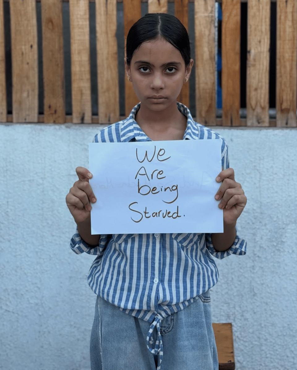 A young, thin Palestinian girl with shadows under her eyes holds a sign written in english. It says "we are being starved."