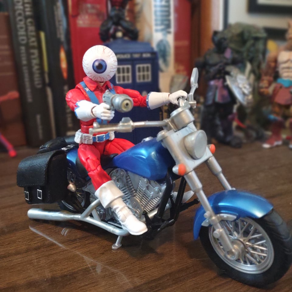 An action figure of a man riding a toy motorcycle. The action figure has a bright red white and blue costume (complete with stars) that looks very much like something a stunt rider would wear in the 70s. Most notably, his helmet looks like a giant white eyeball. The motorcycle is blue with black saddlebags