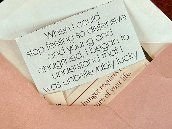 Closeup of a pink on the outside, white on the inside envelope. Coming out of the envelope is a rectangular cut out from a magazine with the quote: “When I could stop feeling so defensive and young and chagrined, I began to understand that I was unbelievably lucky.” Beneath this cut-out is another cut-out with the words “hunger requires” and “of your life.”