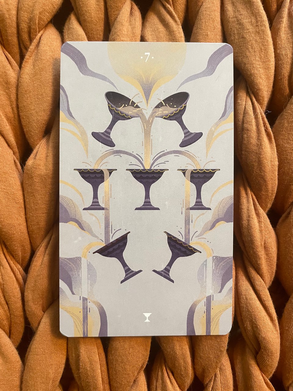 Tarot card featuring seven chalices, with liquid pouring from two upper chalices through three in the middle, and finally into lower ones, set against a pale background with abstract, wavy designs.