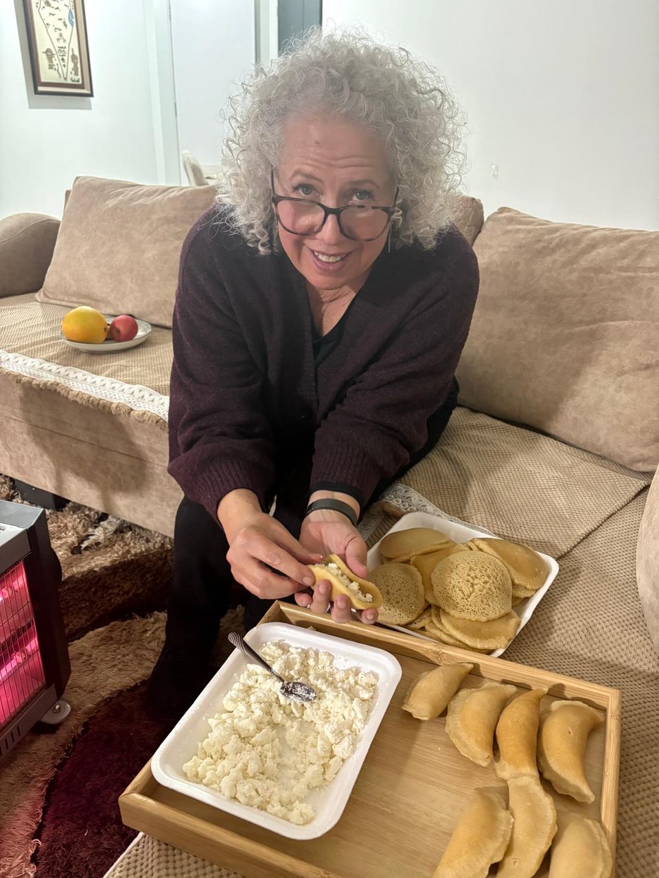 Me sitting on a couch with the ingredients of qatayef (cheese and pancakes), with some of them already stuffed, crimped and ready to be fried
