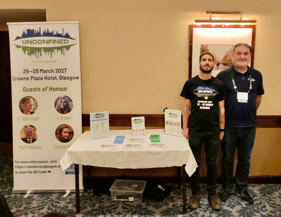 Two men standing to the right of a table with convention leaflets on it, a roll-up banner advertising the convention to the left.