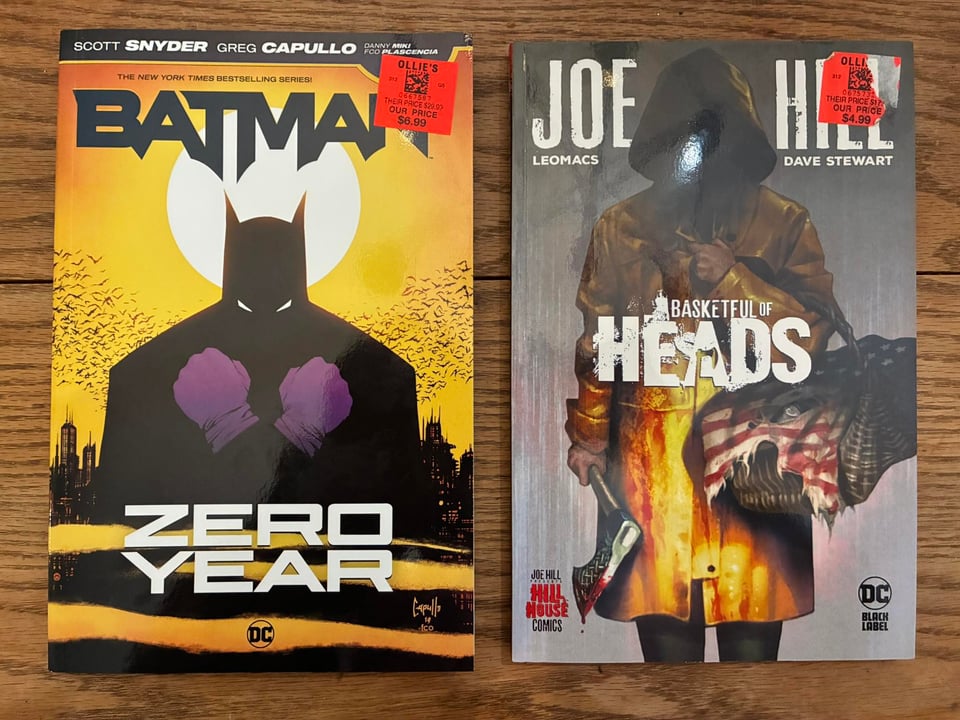 Two books on a wooden table. They are Batman Zero Year and Basketful of Heads. Both of them have orange price stickers on them, which say Ollie's. Both are heavily discounted. Unfortunately, the stickers are very hard to remove.